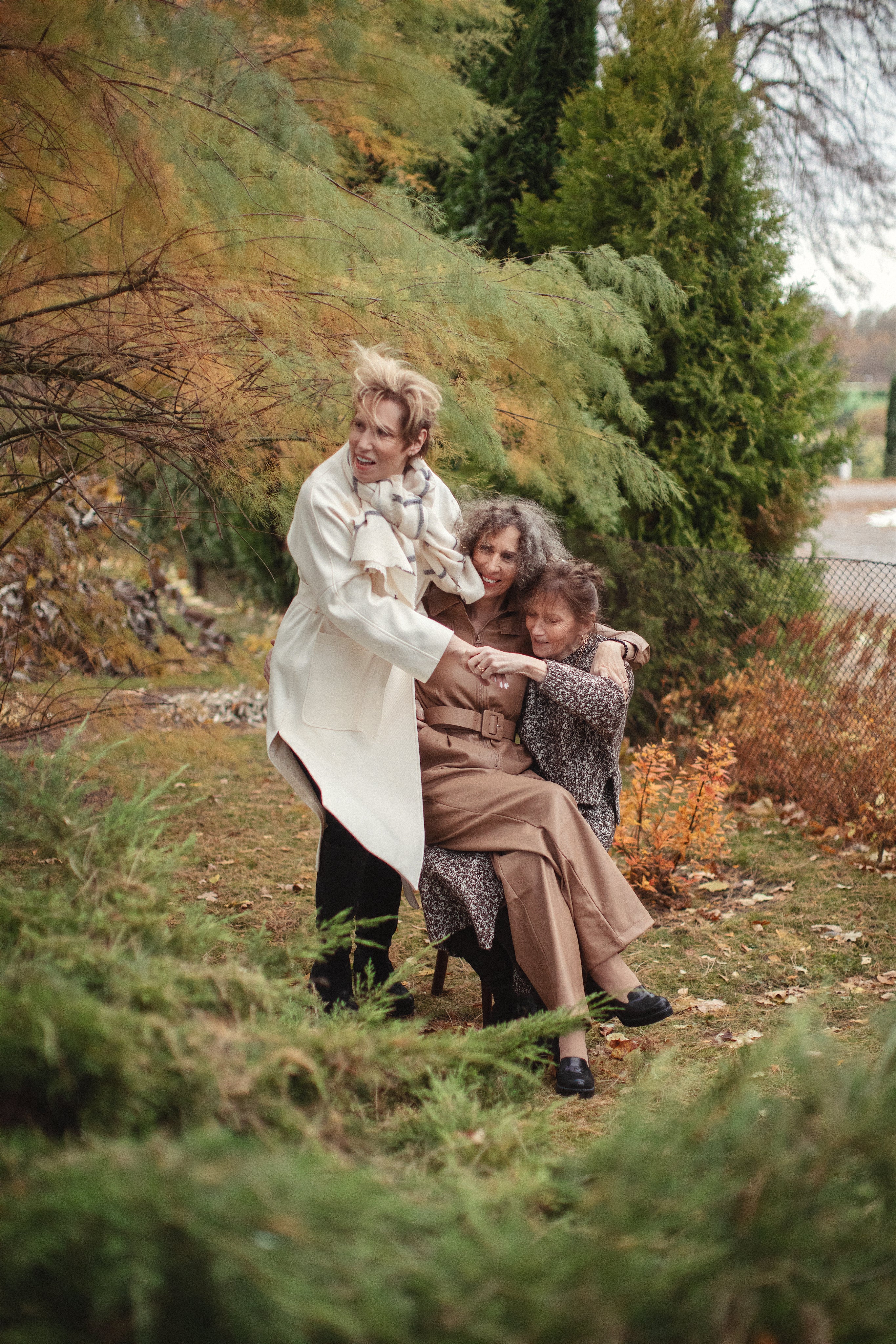 Three women are enjoying time together in a garden. One woman, in a white coat and scarf, stands while the other two women sit. The seated women, one wearing a brown coat and the other a patterned coat, embrace each other. The background has green and autumn-colored foliage.