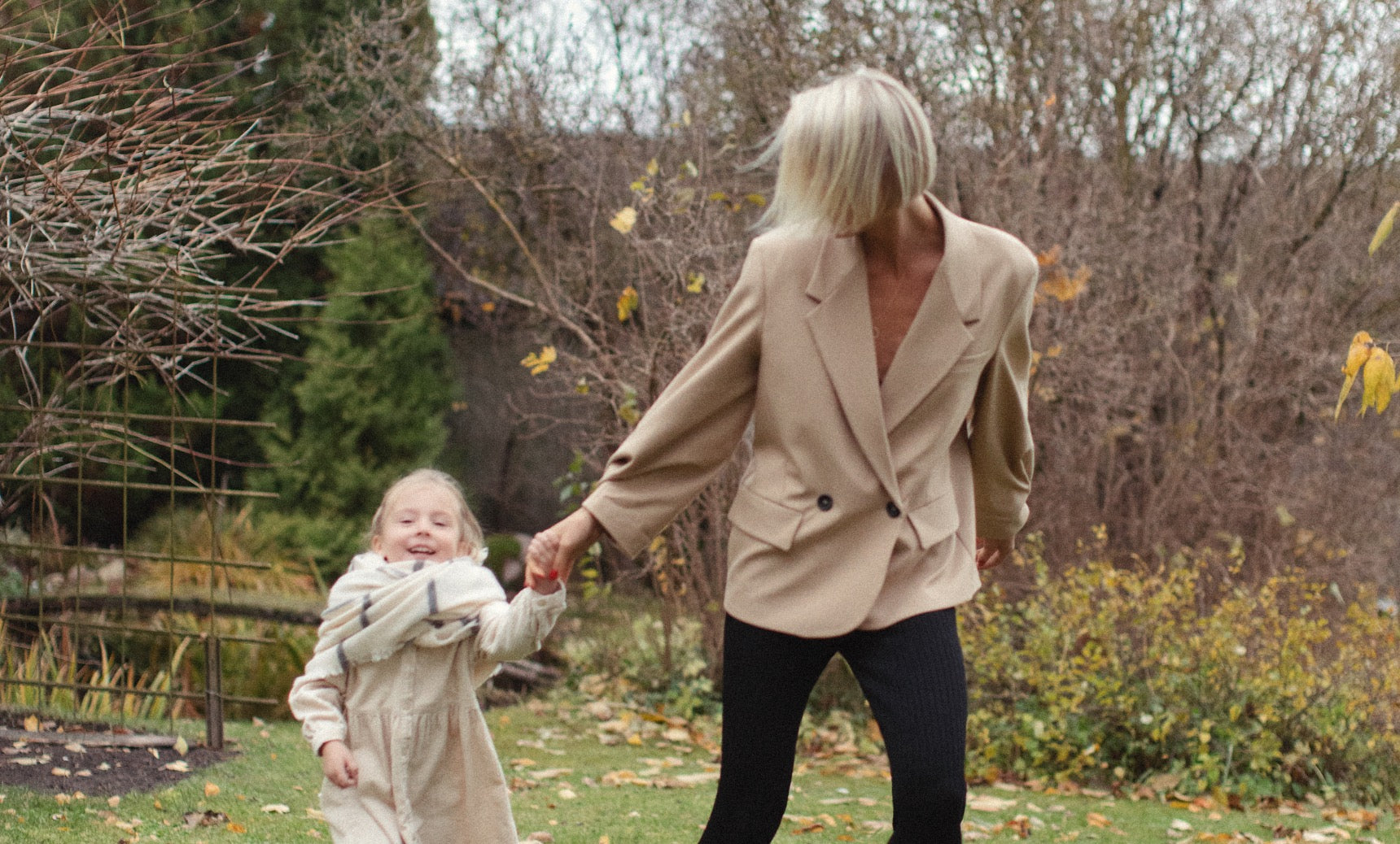 A woman and a young girl run hand-in-hand through a garden. The woman, in a beige blazer and black pants, smiles at the girl, who wears a light-colored dress and a scarf. The background features green foliage and autumn leaves scattered on the ground.