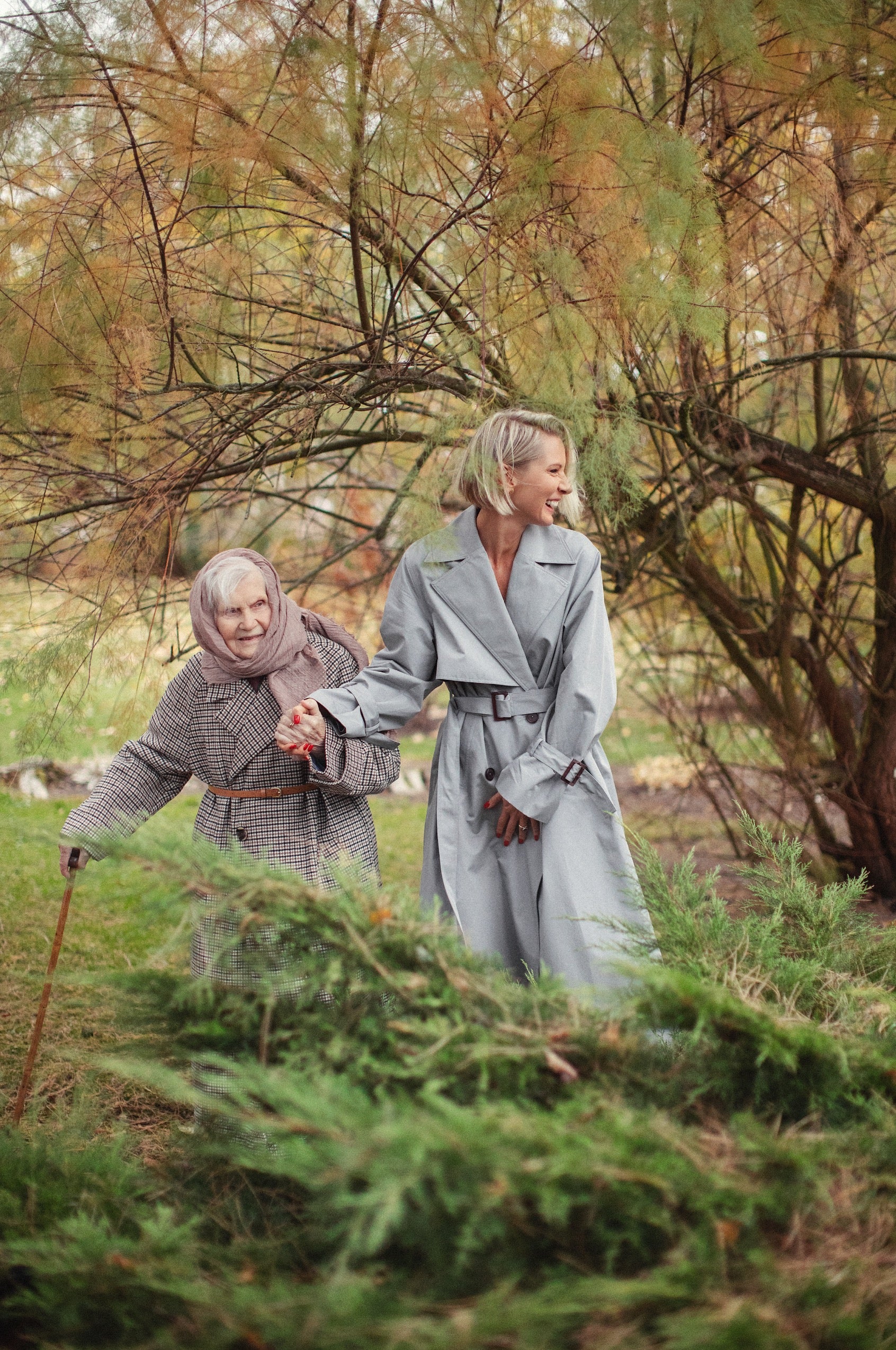 An elderly woman and a younger woman walk hand-in-hand in a garden. The elderly woman, wearing a checked coat and headscarf, uses a cane, while the younger woman, in a light gray trench coat, looks at her and smiles. The background features autumn foliage.
