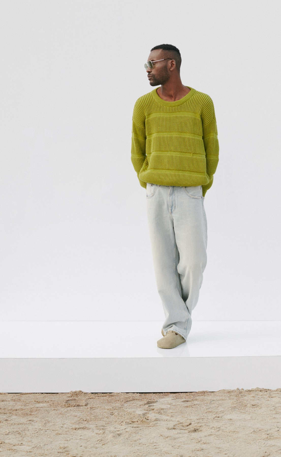 A man stands against a plain white background wearing a lime green knit sweater and light blue jeans. He has his hands in his pockets and wears sunglasses, looking to the side. The scene has a casual and relaxed vibe.